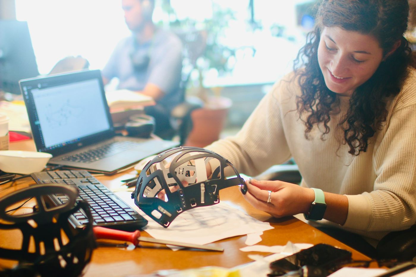 A designer examining a prototype part beside a laptop showing the CAD model
